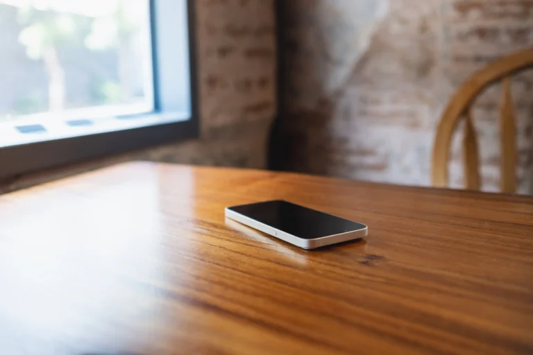 Smartphone with black screen resting on polished wooden table near window and chair.