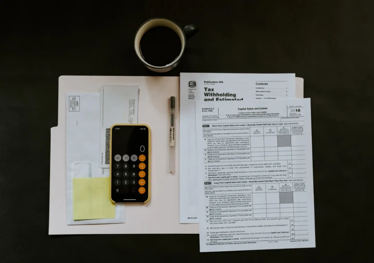 Top-down view of tax documents, calculator on smartphone, pen, coffee cup, and envelopes on dark surface.