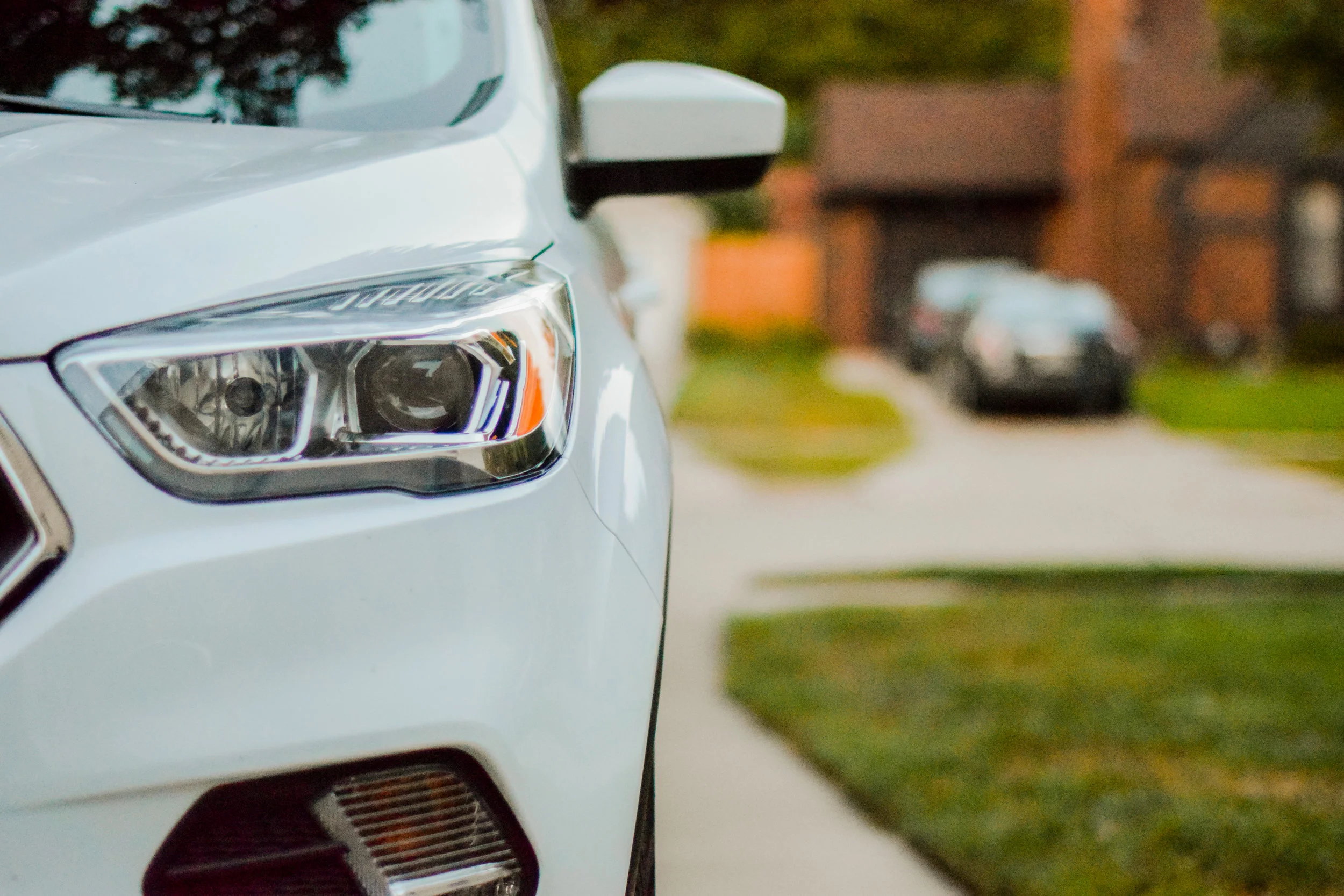 Close-up of a white car's front left headlight and side mirror on a suburban street.