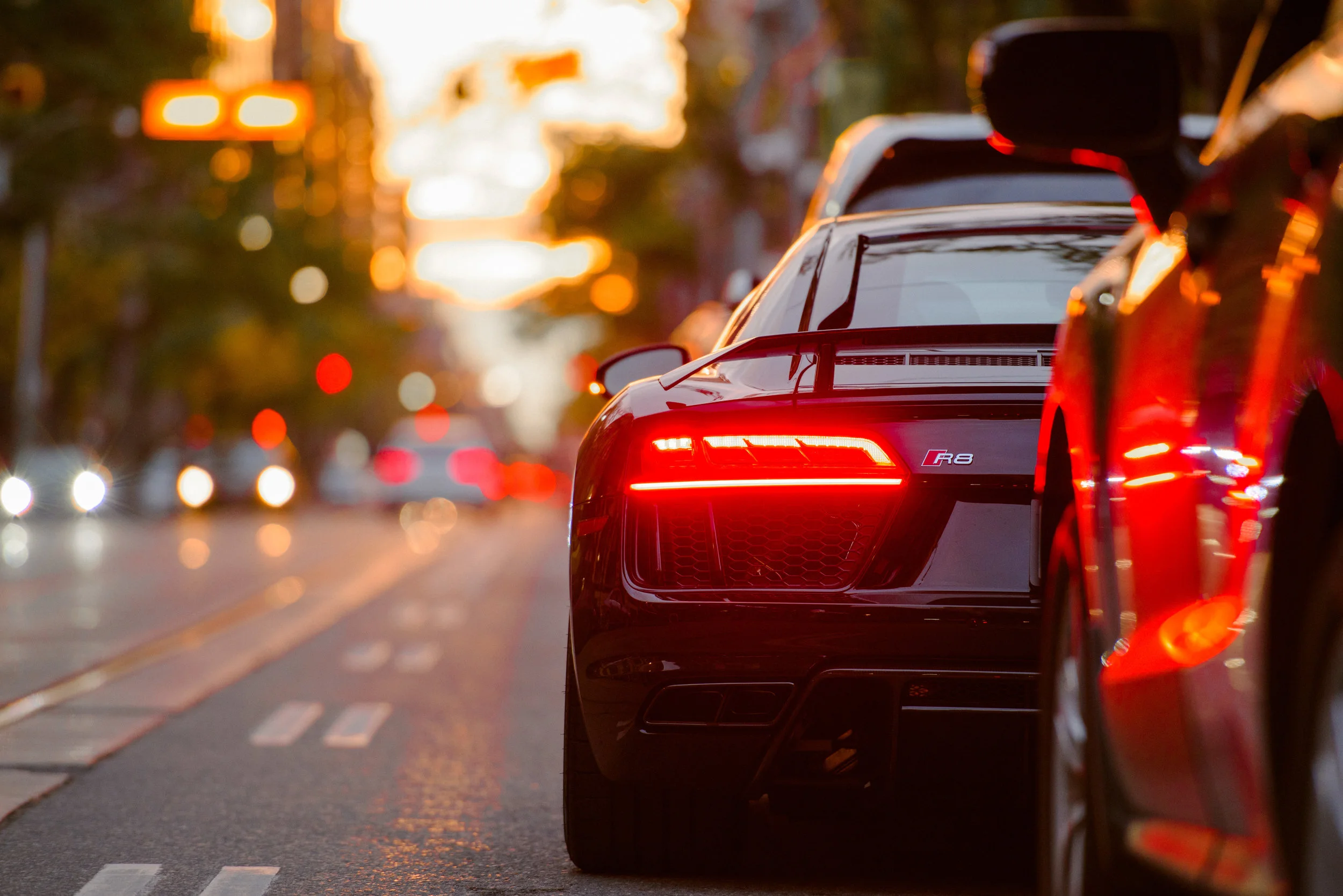 Rear view of a black Audi R8 with illuminated taillights on a city street at sunset.