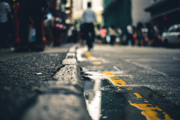 Close-up of a wet street curb with blurred pedestrians and vehicles in the background.