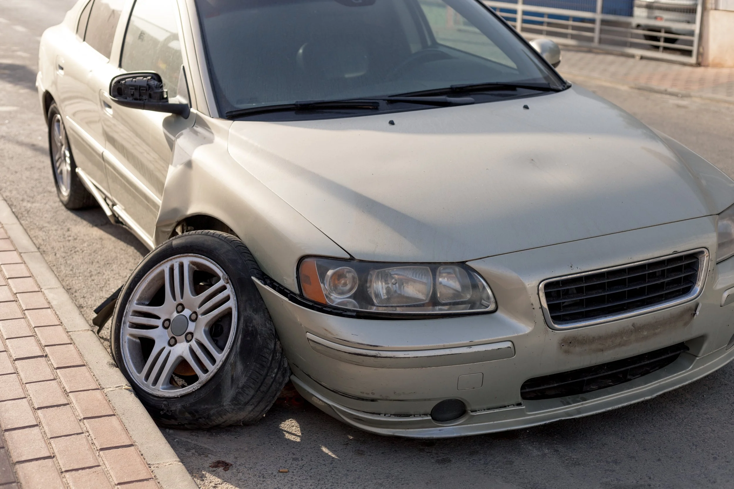 Silver sedan with damaged front left side and detached tire on street curb.