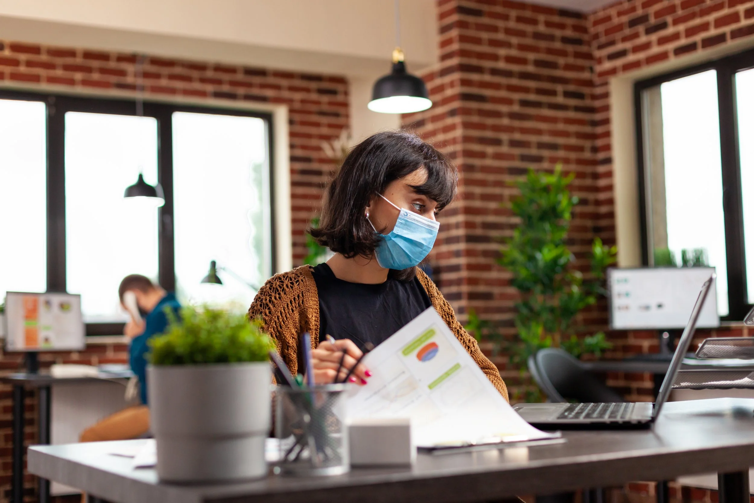 Woman wearing face mask reviewing documents at office desk with laptop.