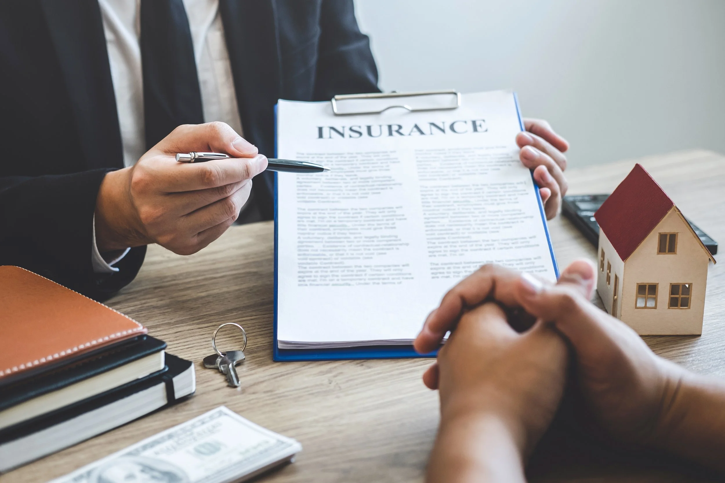 Two individuals discussing a home insurance document with a pen, model house, and keys on table.