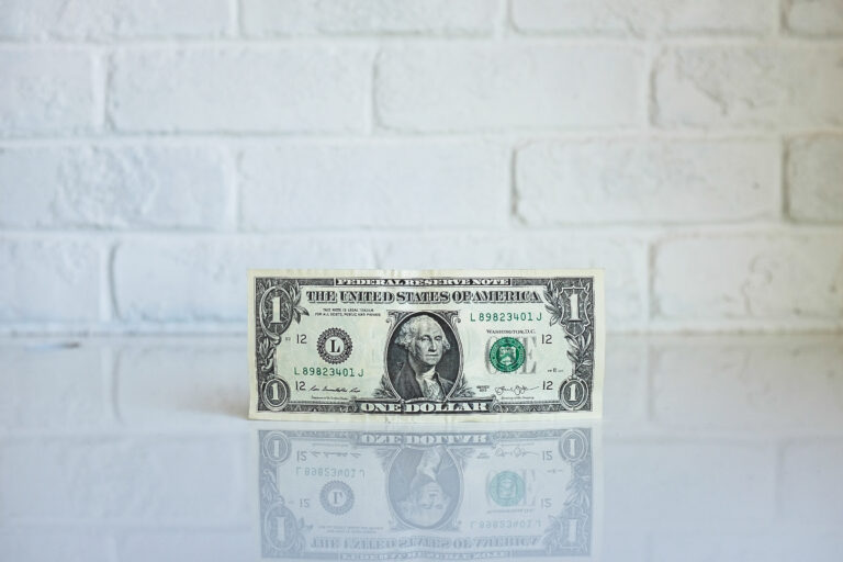 One US dollar bill standing upright on a reflective white surface with a white brick wall background.