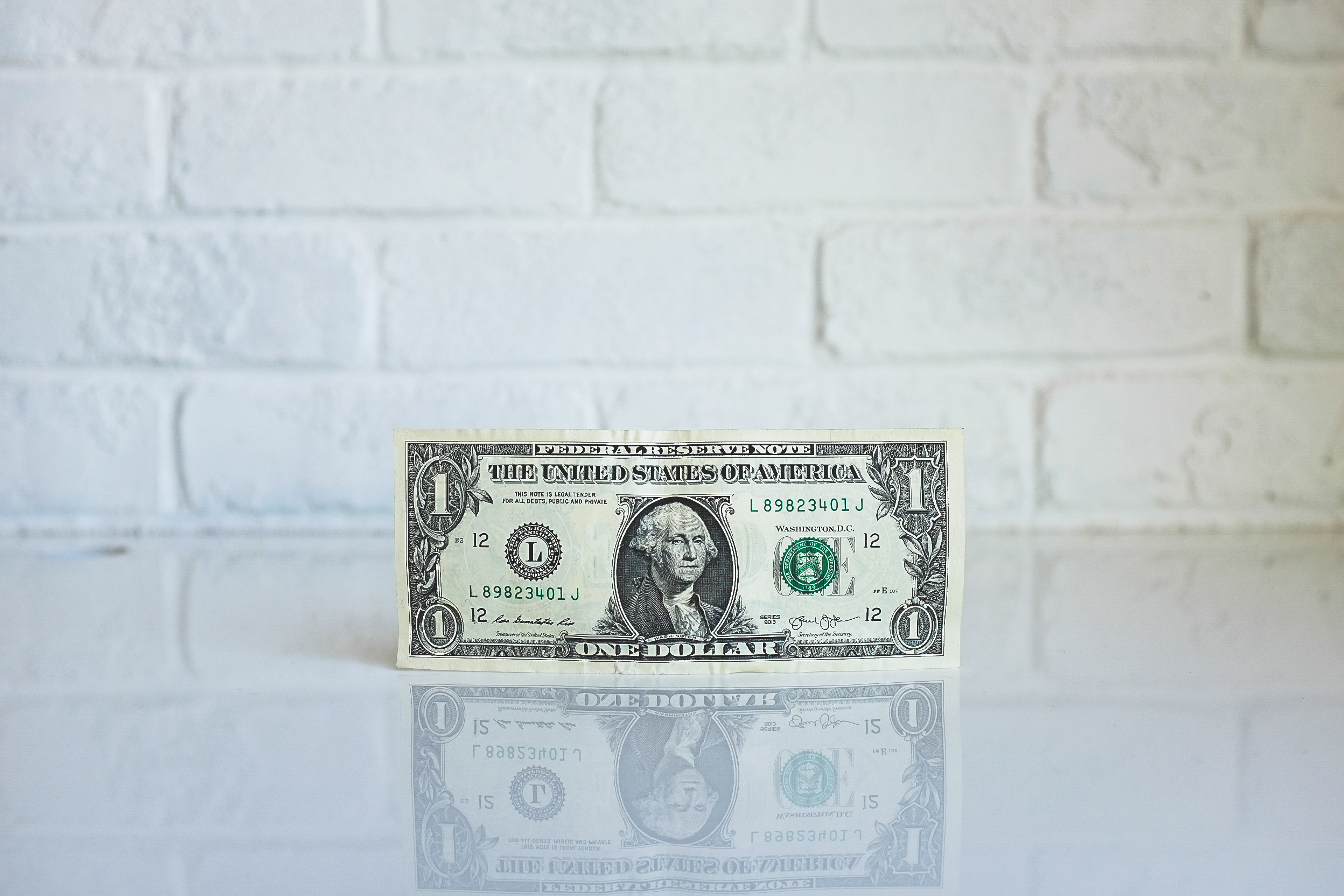 One US dollar bill standing upright on a reflective white surface with a white brick wall background.