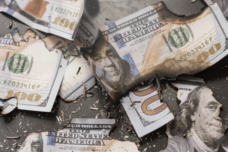 Burnt and torn fragments of United States $100 bills scattered on a dark surface.