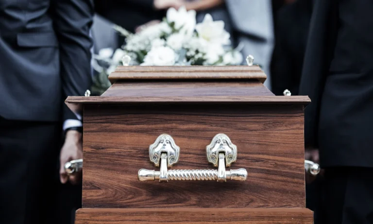 Close-up of a wooden coffin with silver handles carried by people in black suits.