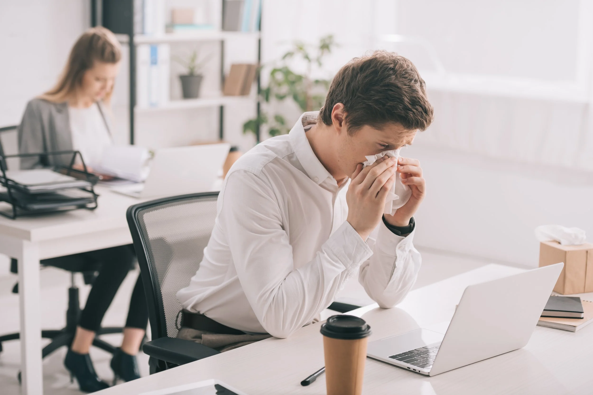 Young man in white shirt wiping nose with tissue at office desk with laptop and coffee cup.