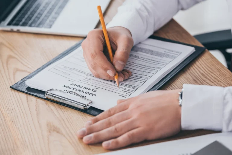 Person in white shirt filling out workers' compensation claim form on clipboard.