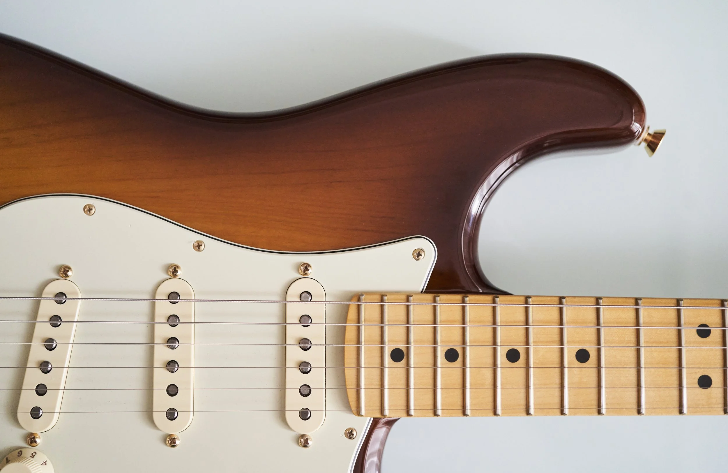 Close-up of a sunburst electric guitar body and maple neck with six strings and white pickguard.