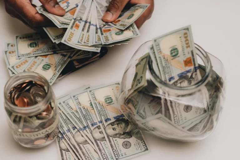Hands holding multiple US $100 bills near a jar filled with cash and coins labeled "Savings.