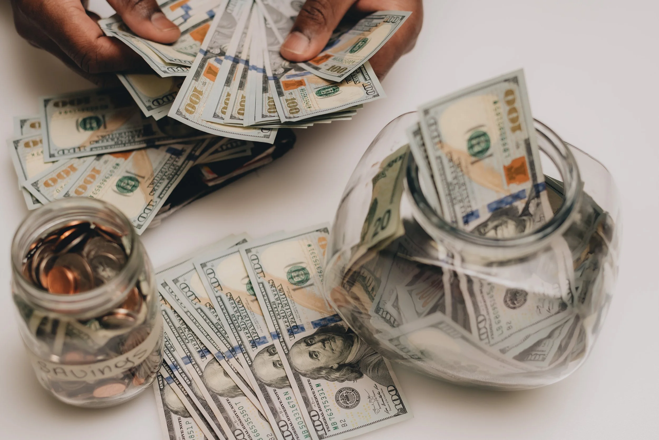 Hands holding multiple US $100 bills near a jar filled with cash and coins labeled "Savings.