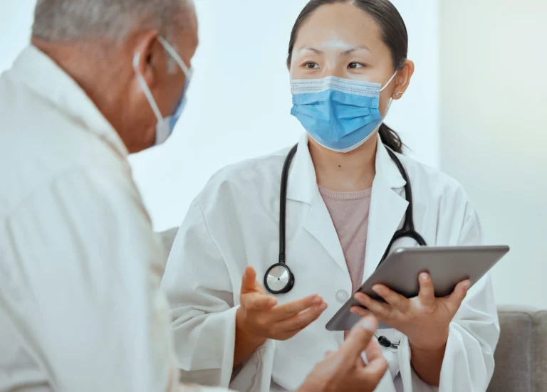 Female doctor wearing a mask and white coat consulting an elderly male patient.