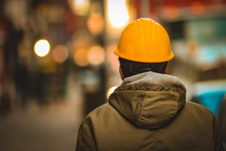 Rear view of a person wearing a yellow hard hat and green jacket in a blurred urban setting.