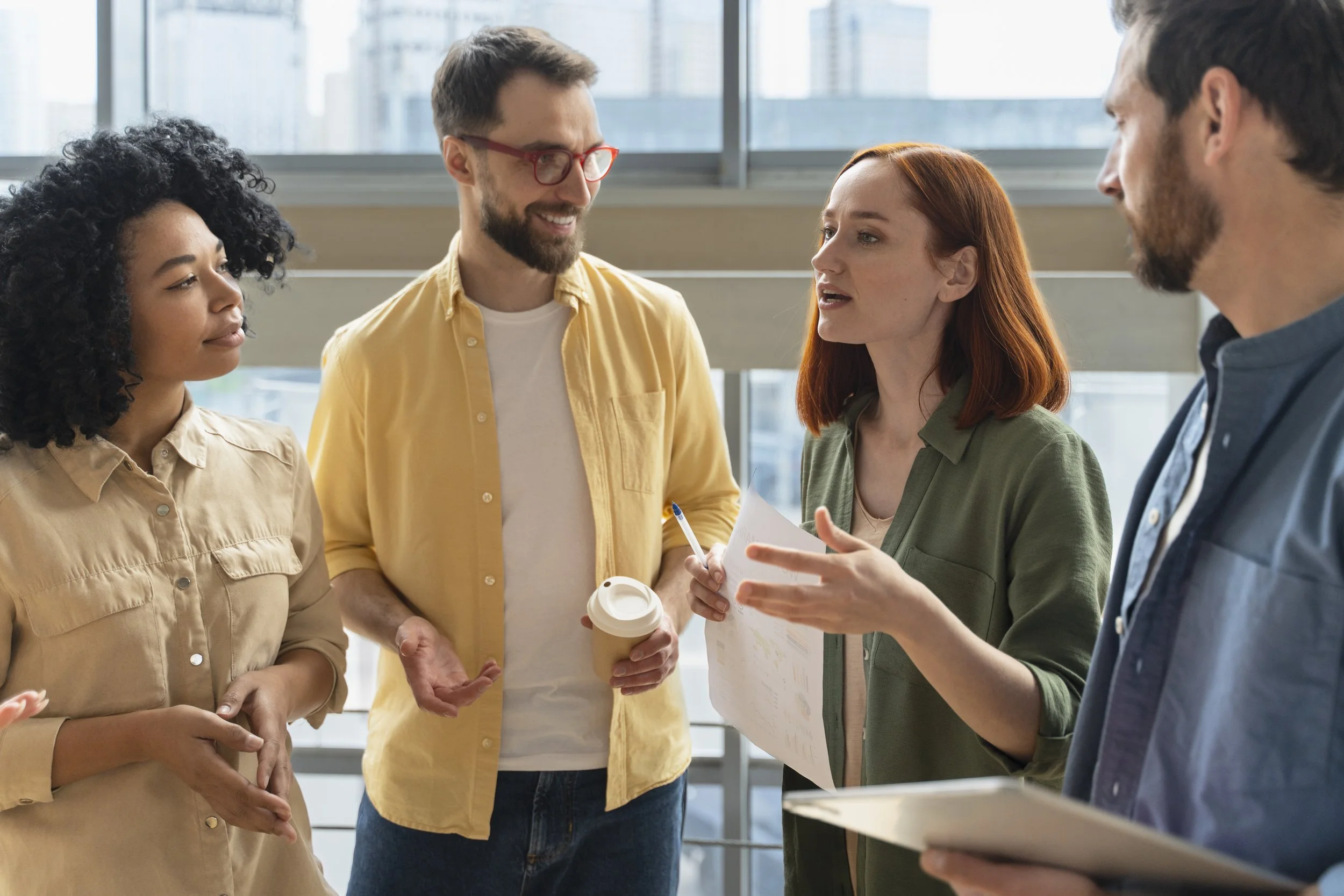 Four diverse professionals engaged in discussion, holding documents and coffee cup, in modern office.