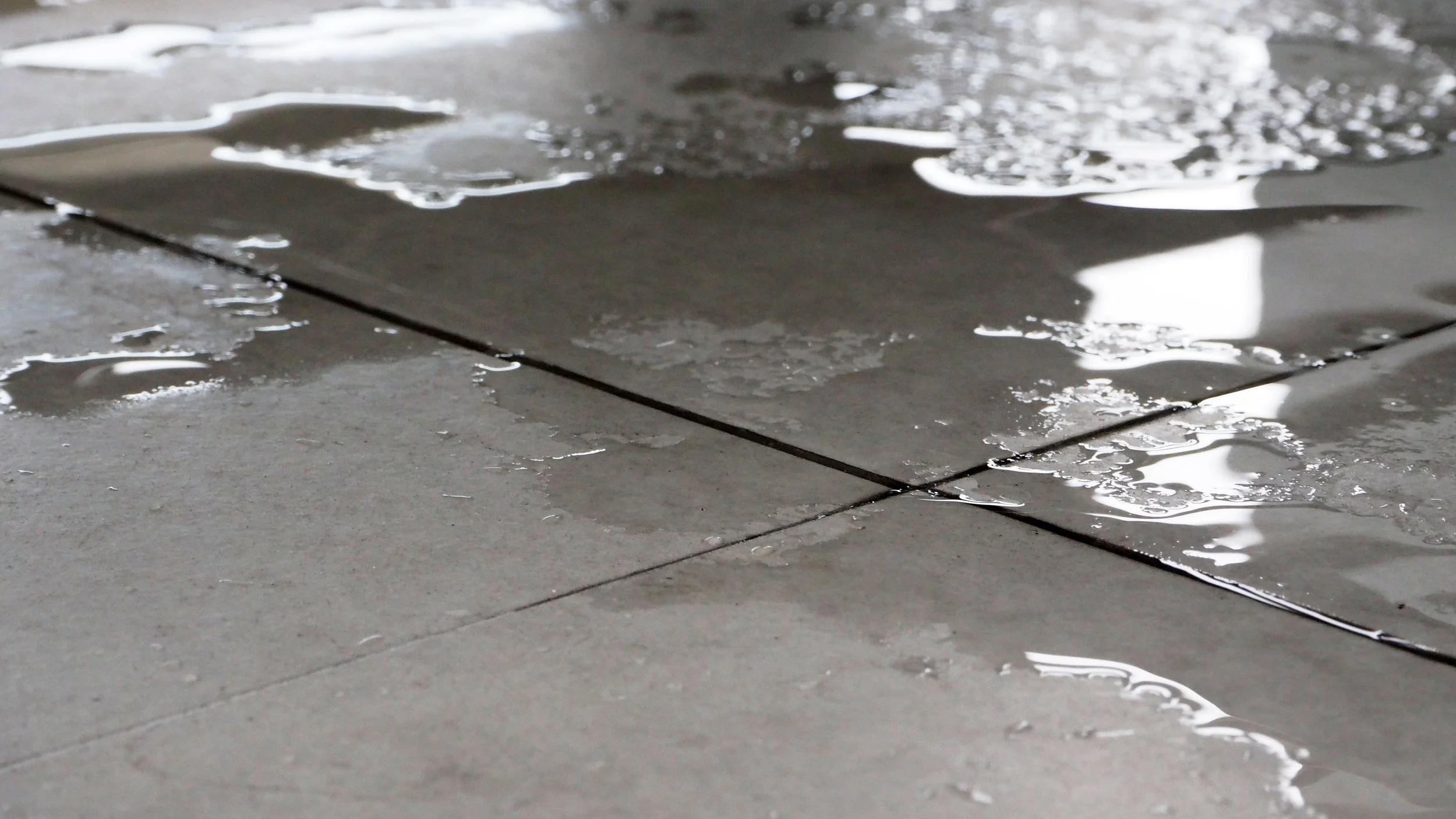 Close-up of gray floor tiles with scattered water puddles and reflections.