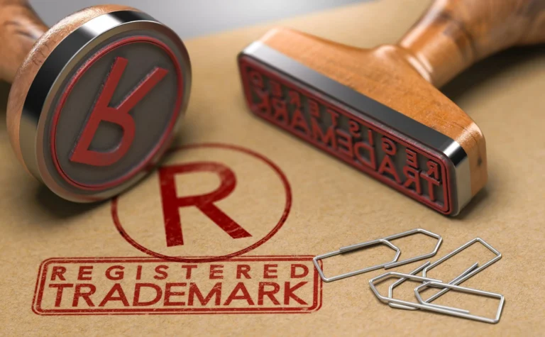Close-up of two wooden rubber stamps, one imprinting "REGISTERED TRADEMARK" in red on brown paper, with scattered silver paperclips.