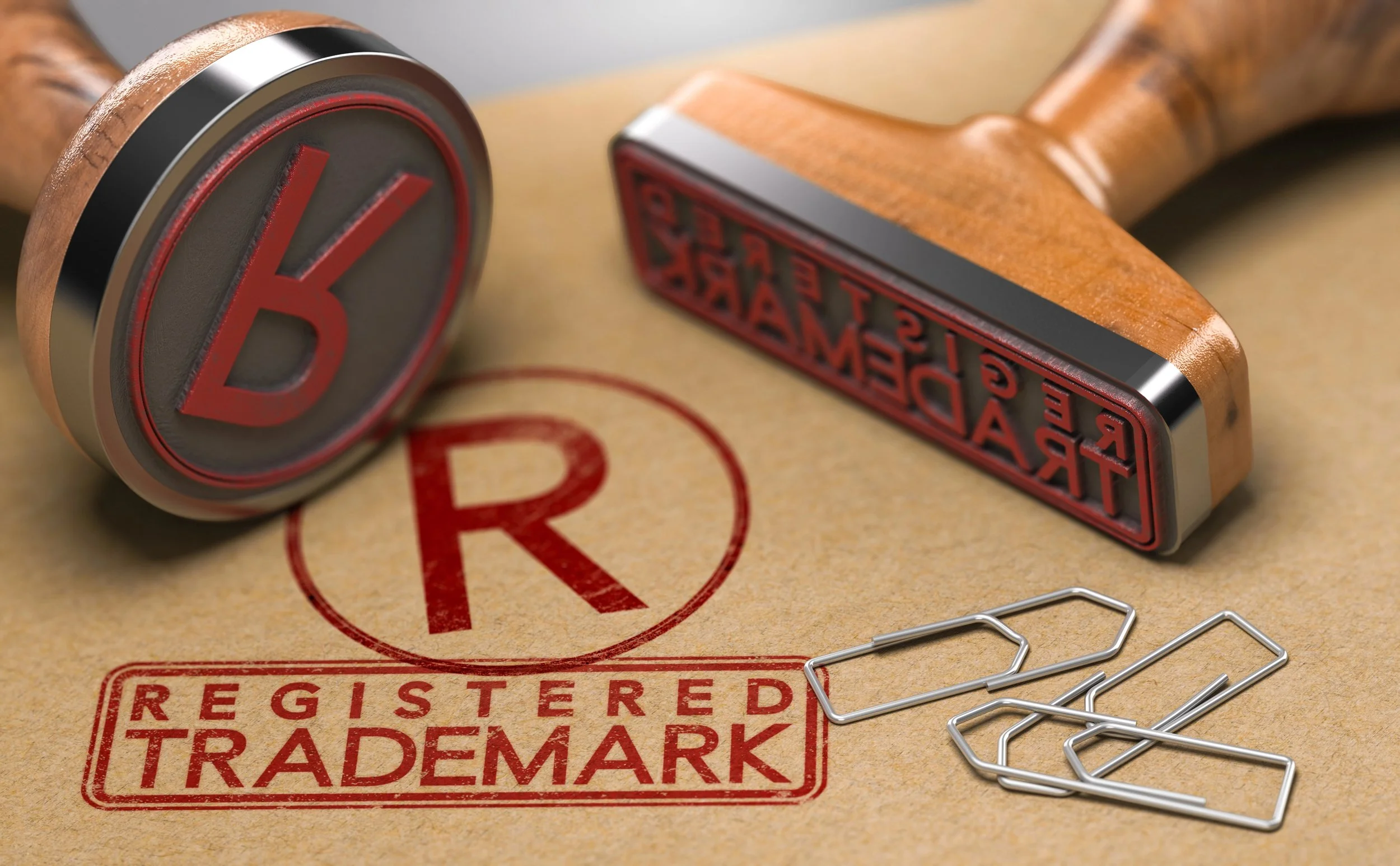 Close-up of two wooden rubber stamps, one imprinting "REGISTERED TRADEMARK" in red on brown paper, with scattered silver paperclips.