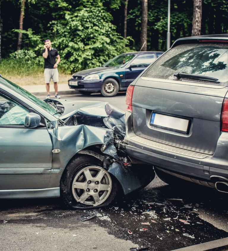 Two silver cars involved in a rear-end collision on a street with scattered debris and a man in the background on a phone.