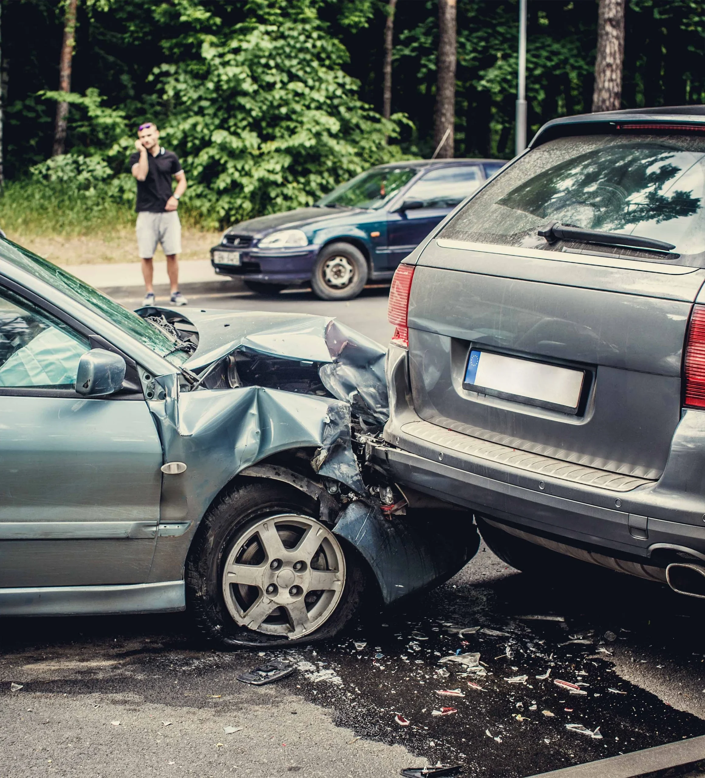 Two silver cars involved in a rear-end collision on a street with scattered debris and a man in the background on a phone.