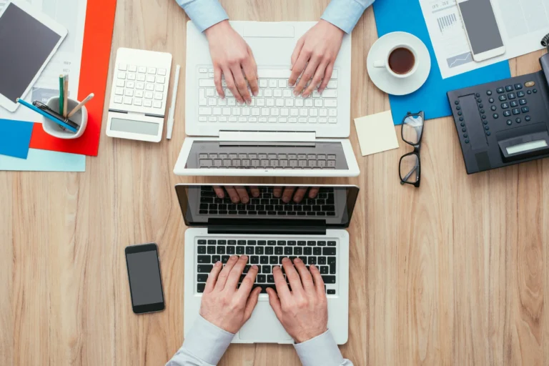 Overhead view of two people typing on laptops amid office supplies and documents.