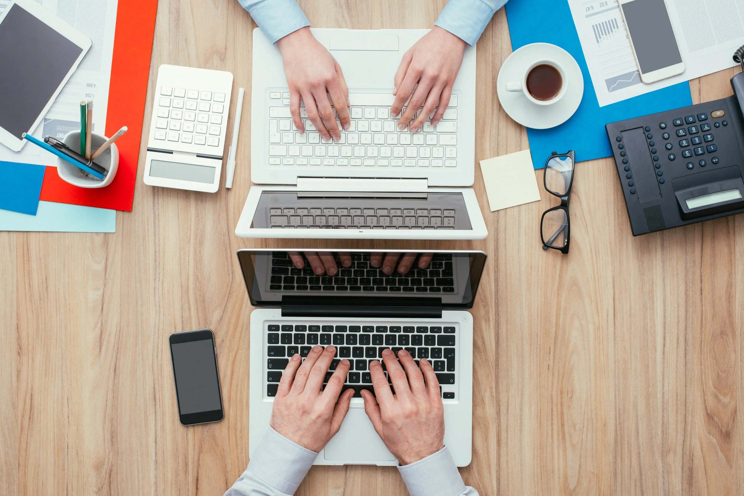 Overhead view of two people typing on laptops amid office supplies and documents.