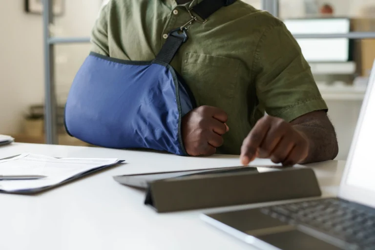 Person with arm in blue sling using a tablet at a desk with documents and laptop.