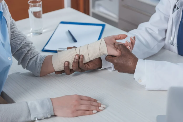 Doctor examining patient's bandaged forearm during medical consultation at desk.