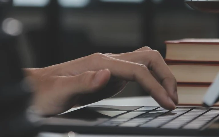 Close-up of hands typing on a laptop keyboard with stacked books in the background.