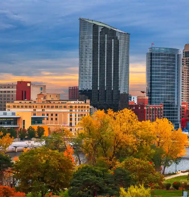 City skyline with modern glass buildings and autumn trees under a cloudy sky.