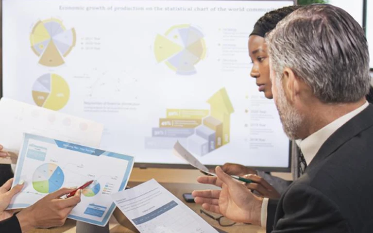Two professionals reviewing printed charts and graphs with a large digital presentation in the background.