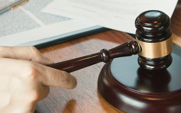 Close-up of a hand holding a dark wooden judge's gavel on a wooden desk with documents.