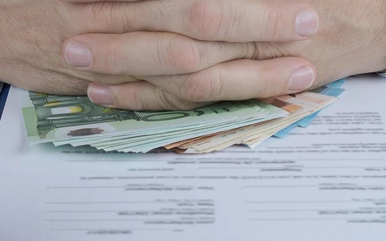 Hands resting on a stack of Euro banknotes placed over a blurred legal document.