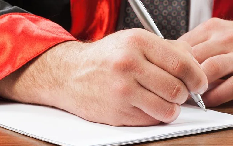 Close-up of a hand in a red academic gown writing with a silver pen on white paper.